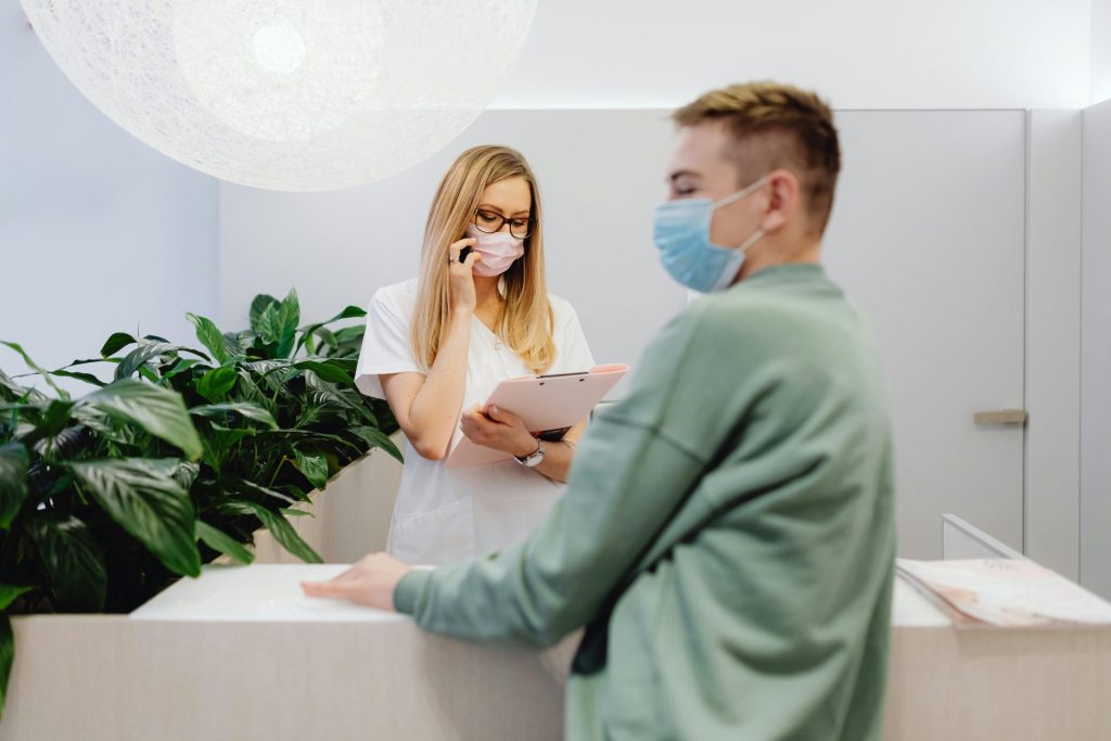 Healthcare worker and patient wearing masks at clinic front desk during pandemic, highlighting safety measures.